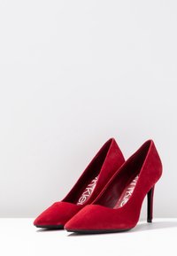 Escarpins hauts en daim rouge avec un bout pointu et un talon aiguille fin, présentant un intérieur lisse et un design minimaliste.