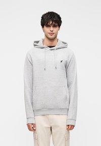 Γκρι φούτερ με κουκούλα και υφή με ανάγλυφο σχέδιο, ρυθμιζόμενα κορδόνια, μπροστινή τσέπη και μικρό μαύρο λογότυπο στο στήθος. Φορεμένο με ανοιχτόχρωμο παντελόνι.