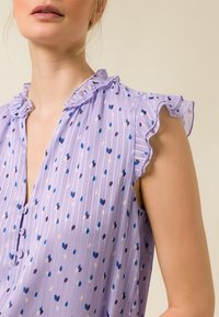 Blusa lavanda con colletto arricciato e maniche a cappuccio, caratterizzata da un motivo di pois multicolori e strisce verticali. Chiusura anteriore con bottoni.