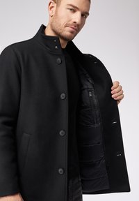 Zwarte jas met een gestructureerd ontwerp, voorzien van een opstaande kraag, zes knopen en een binnenzak met rits. Materiaal lijkt textuurvol en warm.