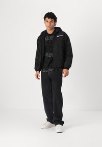 Black hooded jacket with elastic cuffs, worn over a black graphic t-shirt, paired with black jeans and white sneakers. Simple design throughout.