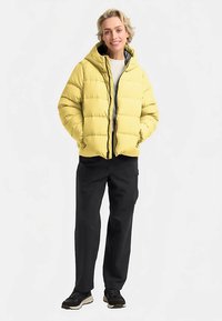 Doudoune jaune avec capuche, fermeture éclair et design matelassé, associée à un pantalon noir et des baskets. T-shirt neutre en dessous.