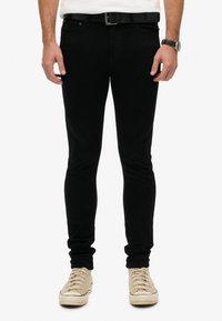 Vaqueros de mezclilla negros con un corte ajustado, que cuentan con cinco bolsillos y una etiqueta de logo sutil. Combinados con zapatillas beige de cordones.