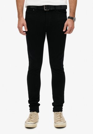 Jeans en denim noir à coupe slim, dotés de cinq poches et d'une étiquette logo discrète. Associés à des baskets beiges à lacets.
