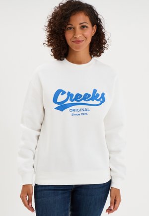 Kobieta o kręconych włosach, ubrana w biały sweter z niebieskim napisem "Creeks Original Since 1974", stojąca na jednolitym białym tle.