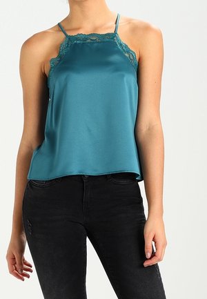 Femme portant un top caraco en satin bleu-vert avec bordure en dentelle et fines bretelles, associé à un jean noir, debout devant un fond uni.