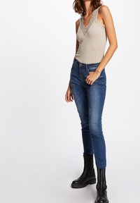 Beige bodystocking med blondedetaljer, parret med blå skinny jeans og sorte ankelstøvler. Glat stof, tætsiddende design, og let glans på bodystockingen.