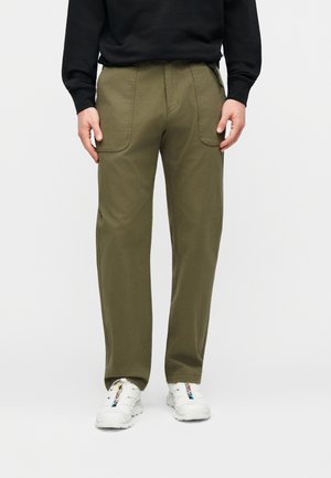 Mand iført olivengrønne bukser med lige ben og forlommer foran samt hvide sneakers, stående mod en ensfarvet baggrund.