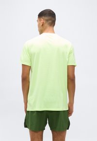 Άνδρας που κοιτάζει προς τα πίσω, φοράει ανοιχτό κίτρινο κοντομάνικο πουκάμισο και σκουροπράσινη βερμούδα, με απλό ανοιχτό φόντο.