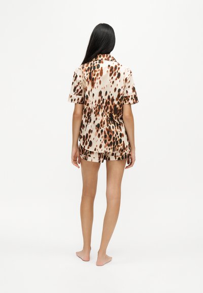 Femme debout pieds nus, de dos, portant une chemise à manches courtes et un short avec un imprimé animal marron et noir sur fond blanc.