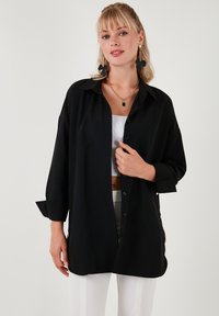 Cămașă neagră, cu nasturi, cu o croială relaxată, mâneci lungi și guler clasic; realizată dintr-un material neted cu un ușor luciu.