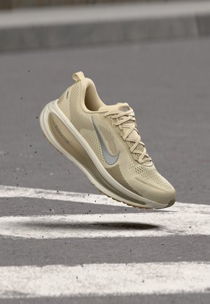 Μπεζ αθλητικό παπούτσι Nike με σόλα ZoomX, κορδόνια και λογότυπο swoosh να αιωρείται πάνω από γκρίζο πλακόστρωτο με λευκές γραμμές.