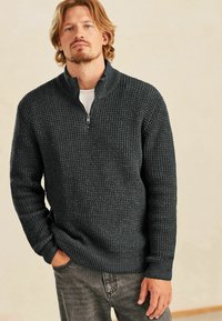 Pulover din tricot gri închis, cu guler cu fermoar de un sfert, model texturat și manșete cu nervuri. Asortat cu blugi gri din denim.