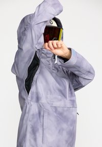 Chaqueta de esquí morada con un material texturizado y resistente al agua. Cuenta con un bolsillo con cremallera y capucha ajustable. Modelo usando gafas de esquí.