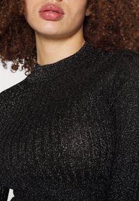 Kvinna med lockigt rött hår som bär en svart glittrig långärmad topp och glansigt rosa läppstift, närbild från halsen till bröstet.