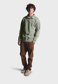Suéter verde oliva con medio cierre y logo en parche, combinado con pantalones marrones y zapatos beige con detalles en azul claro.