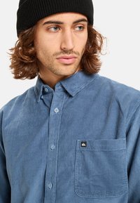 Blauwe corduroyshirt met een button-down kraag, voorzien van een borstzak en een klein logo-label. Zachte textuur en verticale ribbelstructuur zichtbaar.