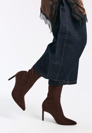 Bruine suède enkellaarsjes met een spitse neus en stilettohak, gecombineerd met donker denim brede broekspijpen. Kantaccent zichtbaar aan de bovenzijde.
