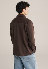 Barna, hosszú ujjú ing, galléros nyakkal, sima textúrával. A hátán lapos yoke és gombos mandzsetta látható, kék farmernadrággal párosítva.