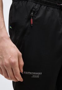 Nærbilde av en hånd som hviler ved siden av svarte Klättermusen Bygvir-bukser med en glidelåslomme og et lite svensk flaggmerke.