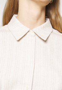 Ingrandimento del collo e della parte superiore del torace di una persona che indossa una camicia color beige chiaro con colletto, caratterizzata da sottili strisce verticali e chiusura con bottoni.