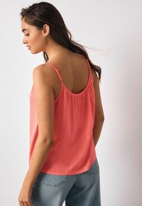 Koralinis berankovis viršus su plonomis reguliuojamomis petnešėlėmis, laisvo silueto, suklostyta nugara, derinamas su šviesiai mėlynais džinsais.