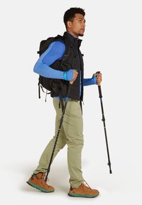 Senderista con chaleco negro, camiseta de manga larga azul, pantalones verde claro y botas de senderismo marrones, sujetando bastones de trekking, caminando hacia adelante.