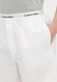 Baltas šorts ar Calvin Klein logotipa elastīgo jostu, ar gludu tekstūru un sānu kabatām, fona ir pelēks tops.
