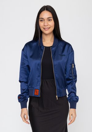 Frau mit langen dunklen Haaren, die eine marineblaue Satin-Bomberjacke über einem schwarzen Kleid trägt und vor einem schlichten hellen Hintergrund steht.