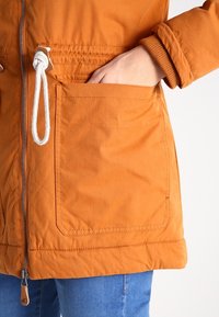 Orange vattenavvisande jacka med dragkedja framtill, elastisk midja och stora patchfickor. Utrustad med dragsko med vit snodd.