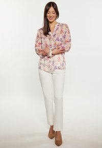 Durchsichtige Bluse mit langen Ärmeln in hellen Pastellfarben, mit floralem Muster und Knopfleiste, kombiniert mit weißen Hosen und beigen High Heels.
