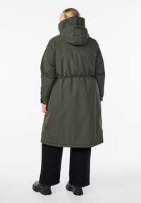 Ženska oblečena v dolg olivno zeleni podložen plašč s kapuco, črne široke hlače in črne škornje, stoji obrnjena stran od kamere na beli ozadju.