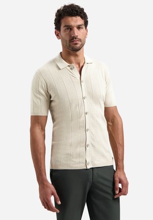 Man met donker krullend haar, gekleed in een crèmekleurig, gebreid overhemd met korte mouwen en knopen, en donkere broek, staand tegen een effen achtergrond.