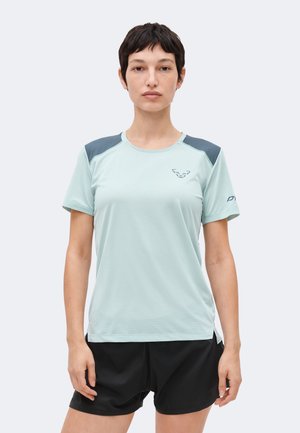 Rövid sötét hajú nő, aki világoskék, rövid ujjú sportpólót visel sötét vállfoltokkal és fekete rövidnadrágot, előre nézve áll.