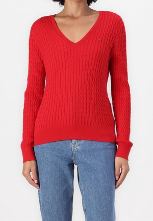 Kobieta ubrana w czerwony sweter z dekoltem w serek i warkoczowym splotem oraz niebieskie dżinsy, pokazana od ramion do bioder na jednolitym tle.