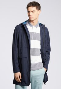 Granatowa kurtka wodoodporna z kapturem, z zamkiem błyskawicznym i kieszenią z przodu, noszona na pasiastej szarej i białej koszulce polo oraz jasnoniebieskich spodniach.