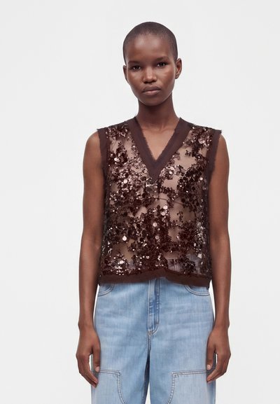 Femme portant un haut marron transparent sans manches à sequins avec un col en V, associé à un jean taille haute bleu clair sur un fond uni.