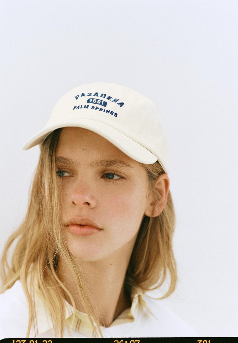 Jeune femme aux longs cheveux blonds portant une casquette blanche avec le texte « Pasadena 1991 Palm Springs », regardant sur le côté sur un fond uni.