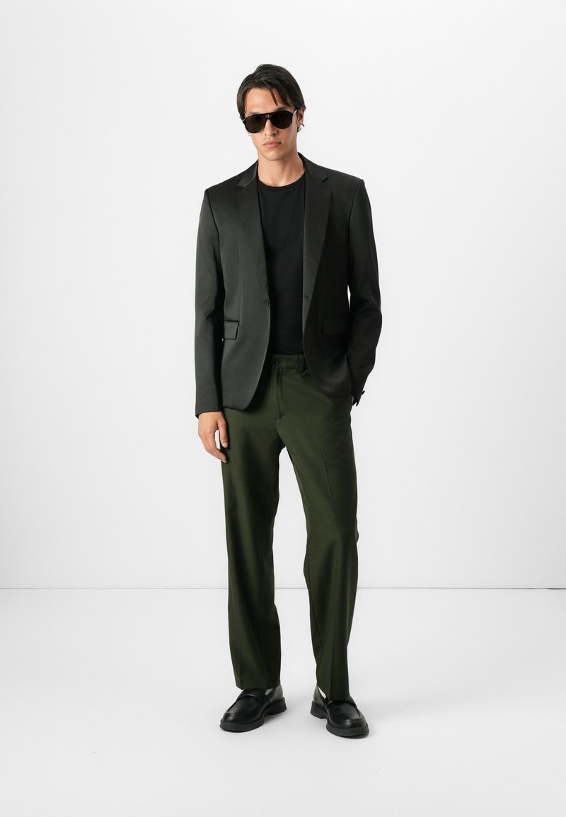 Blazer noir sur une chemise noire, associé à un pantalon vert olive et des chaussures noires. Accessoires comprenant des lunettes de soleil sombres. Look simple et moderne.