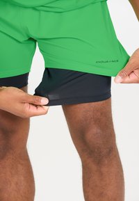 Gröna atletiska shorts försedda med svarta åtsittande innershorts, som visar en lätt och ventilerande design med en slät yta och kontrasterande färger.