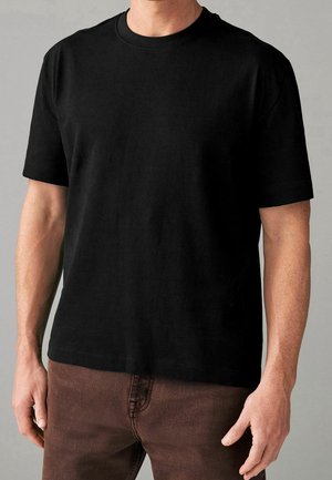 Mand iført en ensfarvet sort kortærmet t-shirt og brune bukser, stående mod en neutral baggrund.