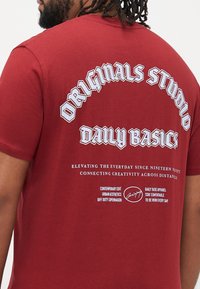 Rød bomulds T-shirt med stort hvidt tekst på ryggen, inklusive "ORIGINALS STUDIO" og "DAILY BASICS," med en glat tekstur og rund hals.
