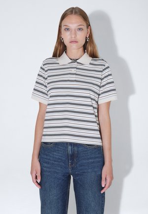Jonge vrouw met lang haar die een wit poloshirt draagt met dunne zwarte en beige strepen, gecombineerd met een blauwe spijkerbroek, staand tegen een witte muur.