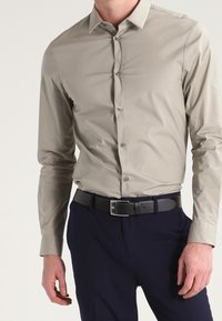 Man i en åtsittande beige knäppskjorta instoppad i marinblå byxor med ett svart bälte, stående mot en enkel bakgrund.