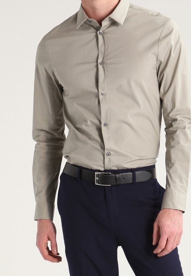 Man i en åtsittande beige knäppskjorta instoppad i marinblå byxor med ett svart bälte, stående mot en enkel bakgrund.