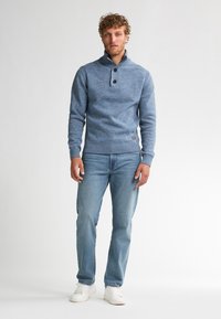 Hombre con cabello rizado que lleva un suéter azul abotonado, jeans azul claro y zapatillas blancas, de pie frente a un fondo blanco liso.
