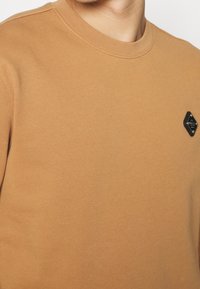 Hnědá bavlněná mikina s kulatým výstřihem a texturovanou černou nášivkou na levé hrudi. Hladká, jednobarevná s ribbovaným výstřihem.