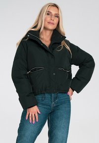 Czarna krótka kurtka puchowa z wysokim kołnierzem, dwoma zamykanymi na zamek kieszeniami z przodu i ściągaczem u dołu. Noszona z niebieskimi dżinsami.