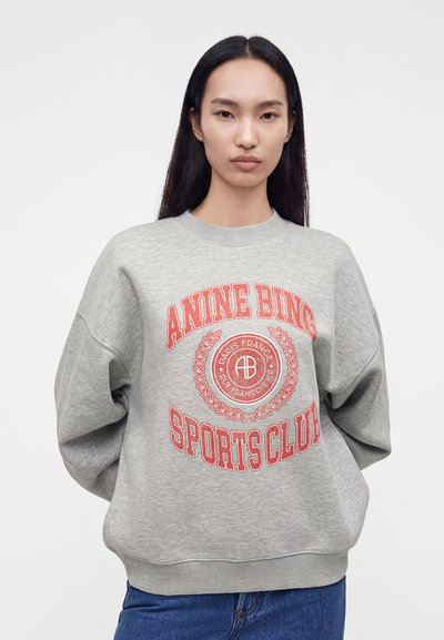 Jeune femme aux longs cheveux foncés porte un sweat-shirt gris avec un texte rouge "ANINE BING SPORTS CLUB" et un jean bleu, debout devant un fond blanc.