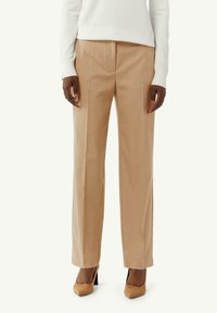 Pantaloni beige su misura con una texture liscia, design a gamba dritta e dettaglio della piega frontale, abbinati a scarpe con tacco alto color marrone.